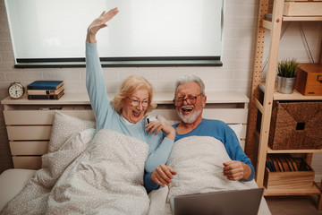 Close photo of elderly couple looking at their smartphones while lying in bed in the morning. Both of them are wearing glasses.