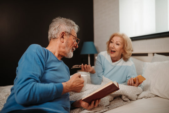 Portrait of lovely senior couple enjoying their time in the morning. Elderly man with classes is holding cup of coffee and reading book while his wife lying next to him and holding smartphone.