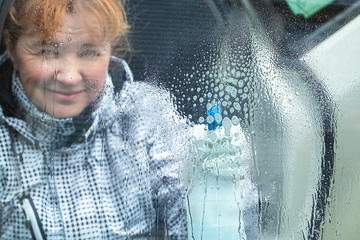 The girl washes a car glass, foam and drops from a spray bottle on the glass. The girl's face is out of focus, blurry. Caucasian with red hair.