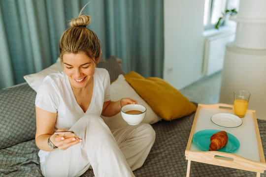 Close Shot Of Woman With Blonde Messy Hair Sending Text Messages From Smartphone While Having Coffee And Breakfast In Bed At Home. Young Woman Decided To Stay At Home Due To COVID19 Pandemic.