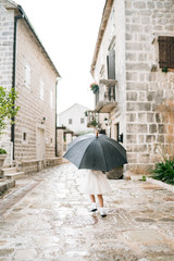 The child poses with an umbrella. A little girl in a dress stands outside under an umbrella black during the rain.
