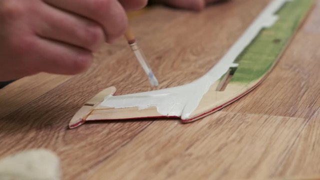 person hand paints wooden outdated passenger aircraft model fin in white with brush on brown table slow motion closeup