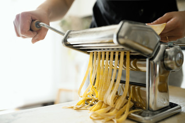Staying at home with your family and preparing fresh home-made pasta (tagliatelle): mom cutting sheets of pasta with pasta machine on a wooden board.