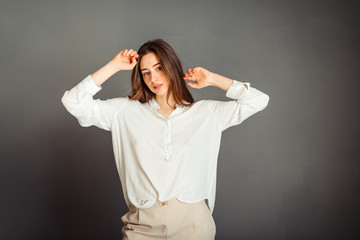 Portrait of a young girl in a white shirt, looking straight, hands near her head bent at the elbows on a gray background. No retouching. Without makeup.