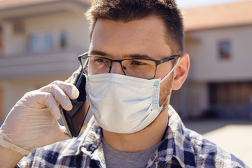 Front view portrait close up on young adult caucasian man in day outdoor wearing protective surgical mask protecting from virus or pollution spread holding mobile smart phone talking and gloves