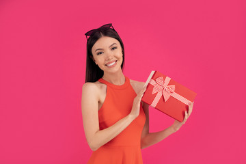 Portrait of a happy young asian woman holding gift box isolated over pink background. dressed in orange dress and sunglasses on head