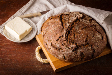 Freshly baked rye bread on wooden board, cotton napkin, butter in butter dish. Wooden background, top view.