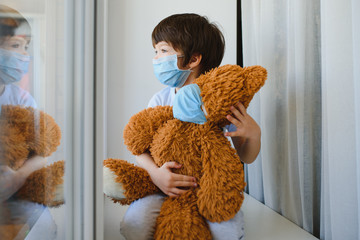 Stay at home quarantine for coronavirus pandemic prevention. Child and his teddy bear both in protective medical masks sits on windowsill and looks out window. View from street. Prevention epidemic.