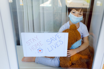 Sad little boy holds a teddy bear looks out the window. Stay at home awareness social media campaign and coronavirus prevention