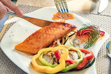 Someone eating salmon filet and vegetables with a fork and knife, on a white plate, restaurant meal, healthy eating