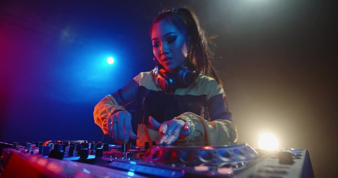 Asian female dj is rocking the party up, creating a mix at turntables in nightclub red and blue lights - nightlife, dance music concept 4k footage