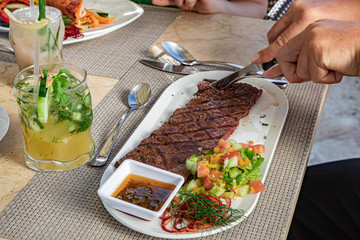 A man eating churrasco beef meat with fork and knife, vegetables and sauce on a white plate, cocktail, restaurant meal