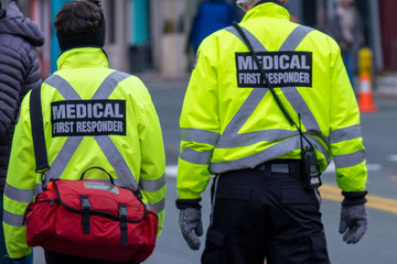 A young medical first responder in a crowd of people walking along a road. She has a red first aid bag over the shoulder of her bright yellow coat or uniform. A tall male responder stands next to her.
