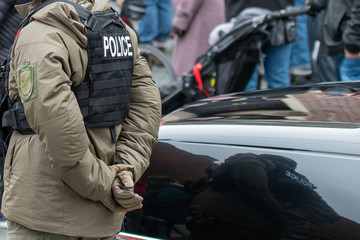 A male police officer wearing a black worn bulletproof vest and a tan colored down filled jacket. The cop is in plain clothes with his hands behind his back.  There are people in the background. 