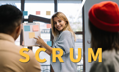 Successful scrum master standing near task board, using sticky notes for productivity. Business colleagues working together, meeting, planning startup, discussion ideas, brainstorming in modern office