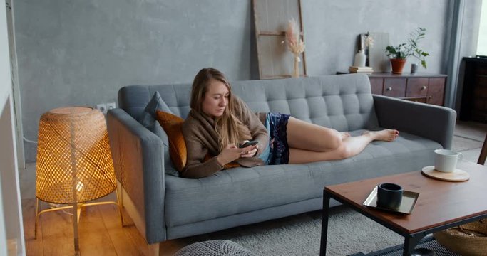 Happy young beautiful Caucasian blonde woman using smart phone at home on cozy sofa, lying down to relax and take a nap.