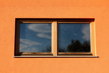 Double windows with dilapidated wooden frame on side wall of suburban family house with reflection of trees and cloudy blue sky on dirty glass on warm sunny autumn day