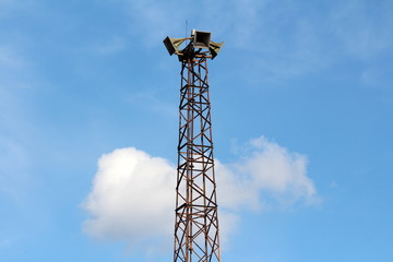 Tall partially rusted metal structure holding four large public civil defence warning air sirens on top on cloudy blue sky background