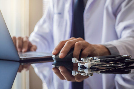 Male doctor in white coat, hand using wireless mouse and work on laptop computer with stethoscope on the desk at office. Meditech, telehealth , online medical, ehr, emr or telemedicine concept. 