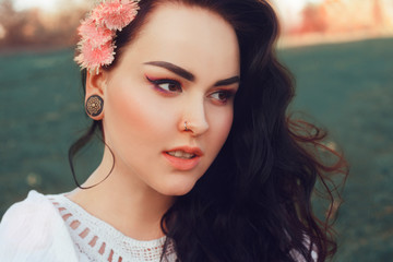 Fototapeta premium Gorgeous brunette sitting in the park on the grass in a white boho dress with pink flowers in her hair in a fashionable finish.