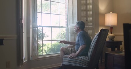 A mature man in his living room gets up from a chair and stands by the window feeling isolated and fearful during the coronavirus pandemic business closures as well as market woes. - Powered by Adobe