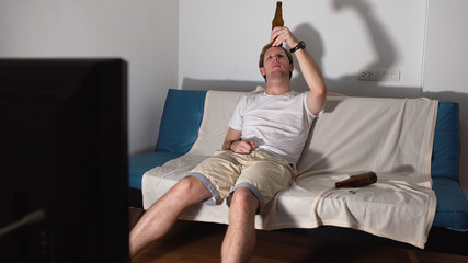 Depressed Young Man Sitting on Couch Drinking Beers While Watching TV Alone