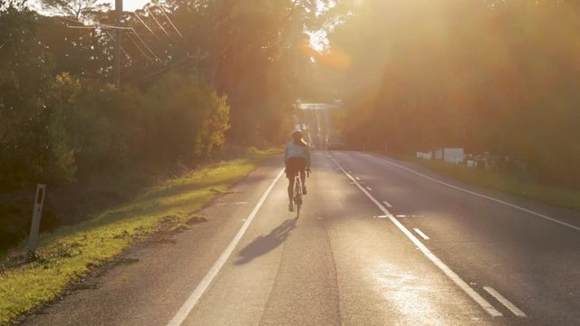 A cyclist rides alone toward sunset/sunrise away from the camera.