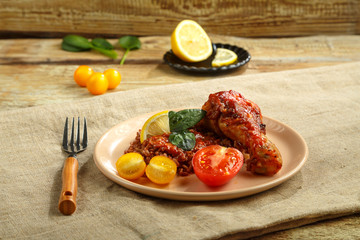 Chicken legs with red rice in tomato-garlic sauce on a plate on a wooden table on a linen napkin and a fork.