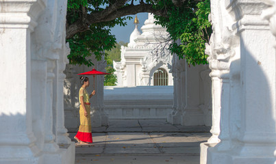 Burmese woman in yellow national costume hold a red umbrella walking at white Kuthodaw  temple in...