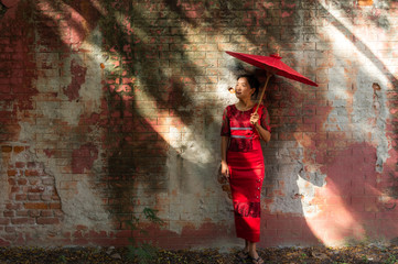 Myanmar woman in red traditional dress holding red umbrella standing at the old wall