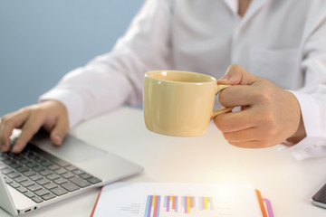 business man in white shirt holding coffee cup and working with computer and document report on desk at home, self quarantine, work from home, marketing, business finance, digital technology concept
