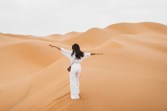 Woman in stylish white shirt and pants with retro photo camera happy to explore Sahara desert sand dunes in Morocco. View from the back.