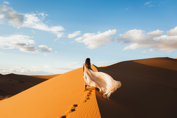 Woman in amazing silk wedding dress with fantastic view of Sahara desert sand dunes in sunset light. Landscape of Morocco, Africa. View from behind. - obrazy, fototapety, plakaty
