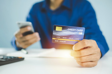 businessman in blue shirt entering security code with mobile phone and paying with mock up credit card and calculator on desk at home, shopping online, electronic pay and digital technology concept