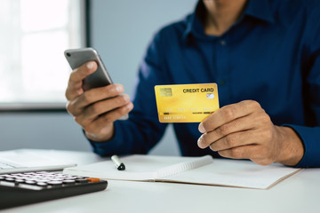 young man in blue shirt entering security code with mobile phone and paying with mock up credit card and calculator on desk at home, shopping online, electronic pay and digital technology concept