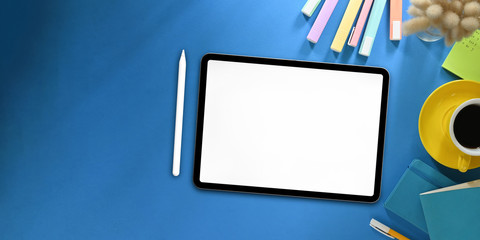 Top view image of white blanks screen computer tablet with stylus pen putting on colorful working desk and surrounded by marker pens, wild grass, coffee cup, note and pen. Orderly workspace concept.