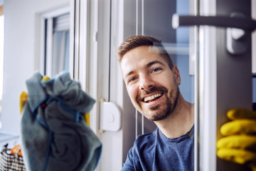 Portrait of young worthy smiling bearded man with rubber gloves on cleaning window at home.