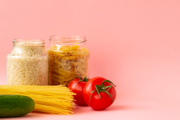 Noodles, rice, pasta in glass jars stand on a pink background. Nearby are red tomatoes and green cucumber. Raw materials for cooking.
