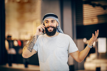 Attractive smiling tattooed bearded arab guy standing indoors and having conversation on smart phone.