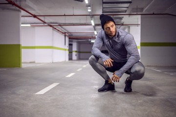 Young muscular man crouching in underground garage and taking a break after exercises.