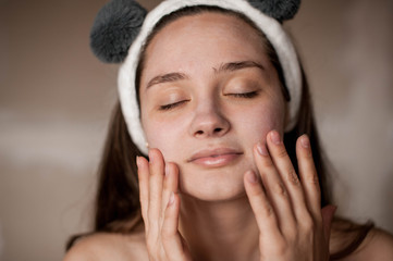 Fototapeta na wymiar a brunette girl without makeup with problematic skin with a bandage with ears on her head naked wrapped in a towel after a bath doing Spa treatments applies a white clay mask to the face