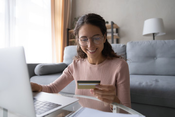 Smiling young female client girl holding credit debit card, shopping securely online on computer from home. Happy millennial woman in eyewear entering data, purchasing goods from internet store.