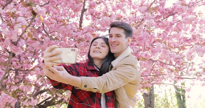Young couple walking the blooming alley between blossom cherry trees at backlit. Happy woman in red coat and man in beige jacket making photos oh themselves on frontal camera