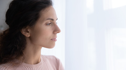 Side close up head shot view thoughtful young woman looking away through window, suffering from loneliness indoors. Attractive millennial lady thinking of problems, staying shelter alone at home.