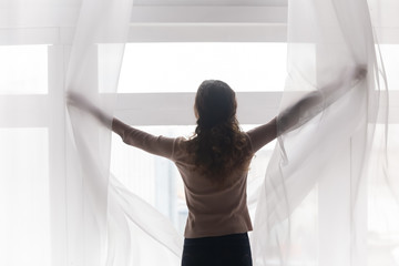 Back rear view happy young woman opening light curtains, welcoming new day. Millennial brunette...