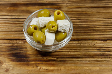 Feta cheese cubes with green olives in glass bowl on a wooden table