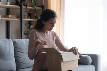 Stressed frowning young woman looking inside cardboard box, feeling dissatisfied with order crashed...