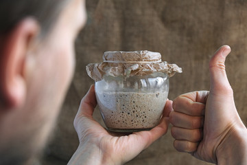 baker hand holds glass jar with sourdough and make thumb up gesture, like sign, brown background,...