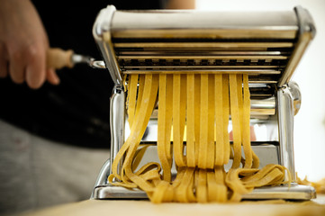 Staying at home with your family and preparing fresh home-made pasta (tagliatelle): mom cutting sheets of pasta with pasta machine on a wooden board.
