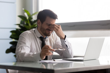 Naklejka premium Stressful businessman taking off glasses, suffering from dry eyes syndrome after long laptop use. Exhausted employee executive manager ceo feeling eye strain, massaging nose bridge